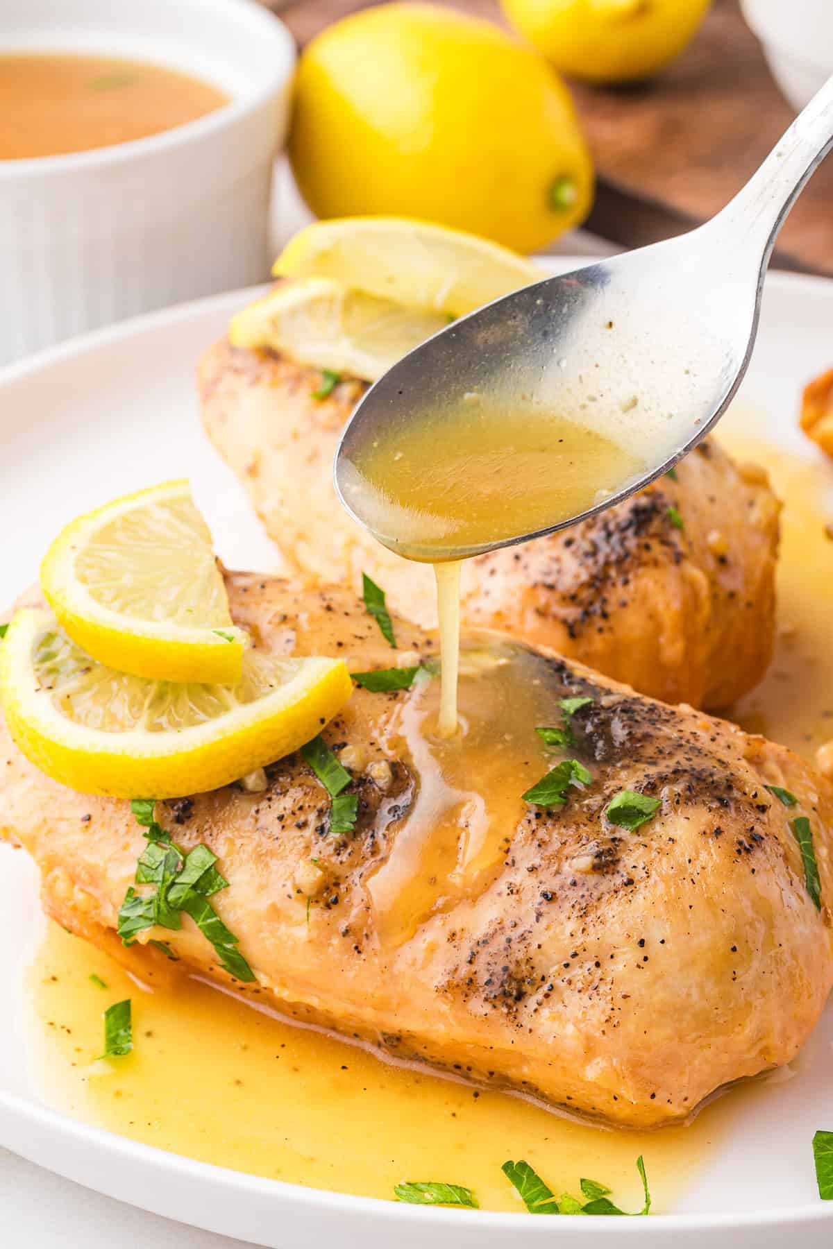 A spoon drizzles lemon sauce over seasoned Lemonade Chicken, garnished with lemon slices and herbs. This delightful chicken recipe is complemented by lemons and a sauce bowl in the background.