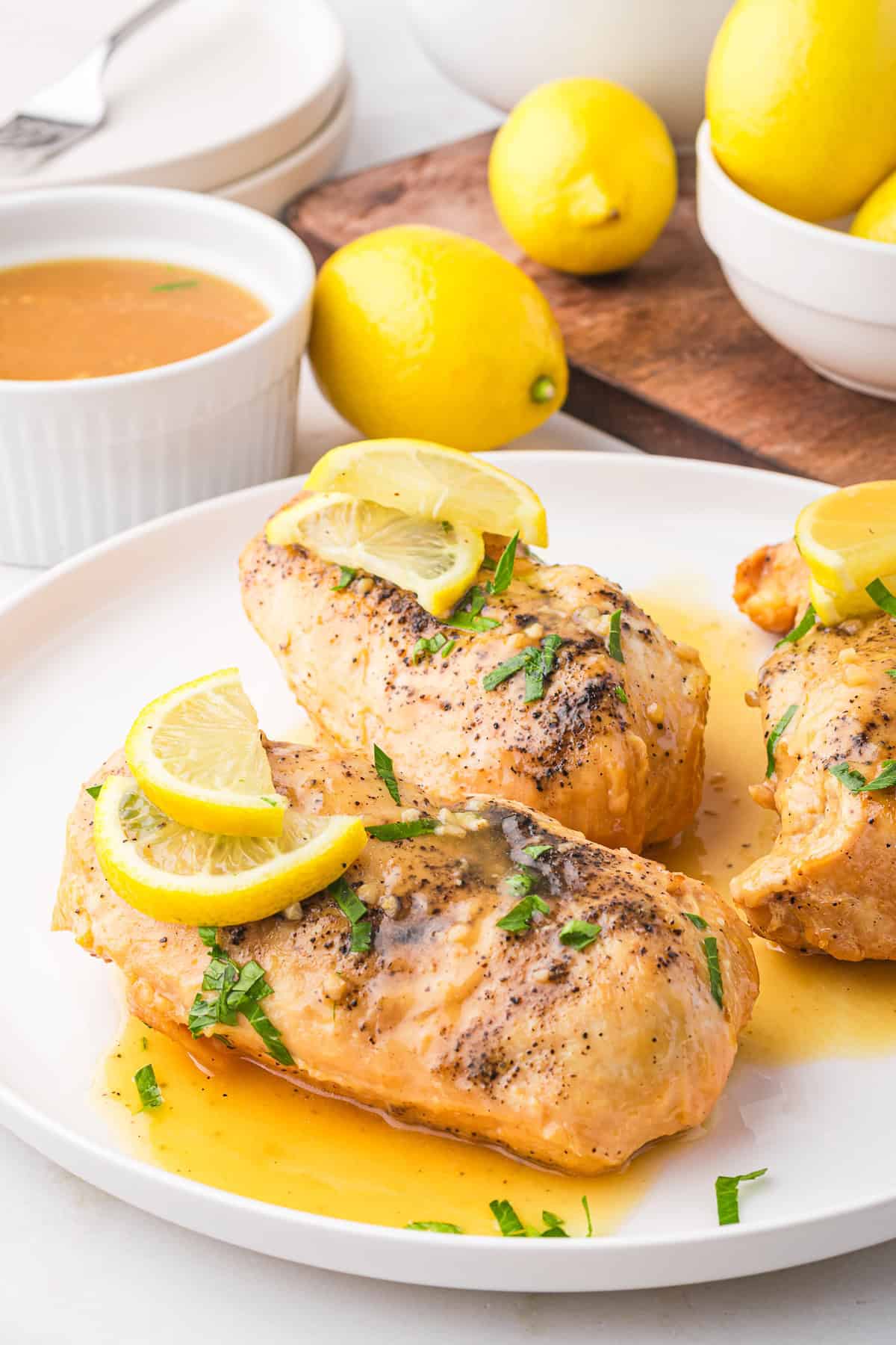 Chicken breasts with lemon slices, garnished with herbs, rest on a plate with sauce, echoing a delicious lemonade chicken vibe. Lemons and sauce linger enticingly in the background.