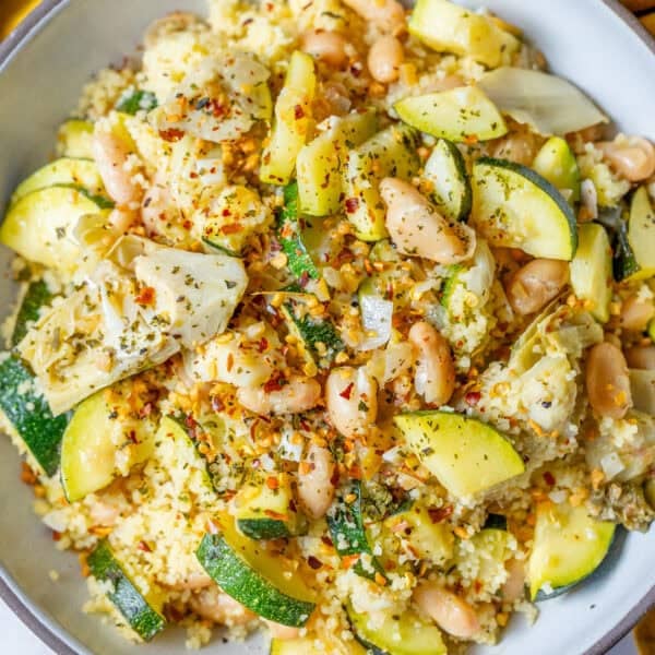 A bowl of couscous mixed with zucchini, white beans, artichoke hearts, and herbs, garnished with red pepper flakes.