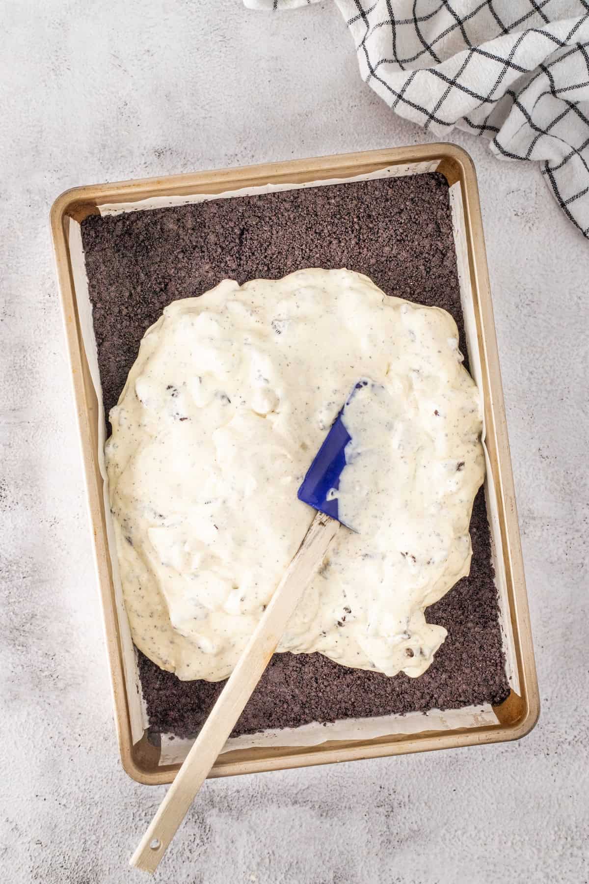 A baking tray holds a layer of crushed cookies beneath a rich spread of cream mixture, perfect for making cookies and cream cheesecake bars. A spatula rests on top, with a checkered cloth placed nearby.
