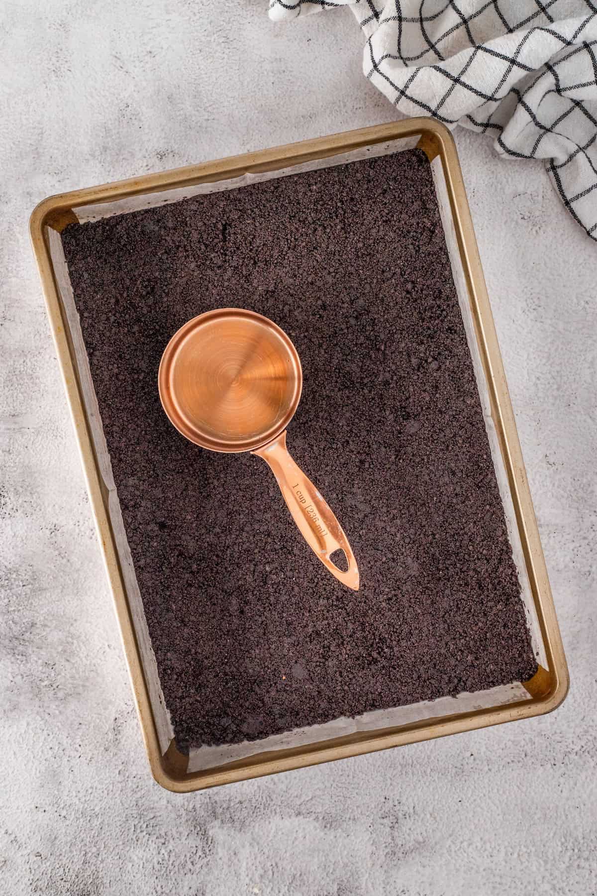 A copper measuring cup rests on a baking tray filled with finely ground dark material, reminiscent of cookie crumbs. A checkered cloth is visible in the top corner.