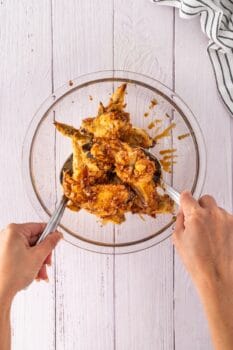 Two hands mix honey-glazed chicken wings in a glass bowl with metal tongs on a white wooden surface. A striped cloth is partially visible in the top right corner, hinting at the tantalizing aroma of garlic mingling with marinated flavors.