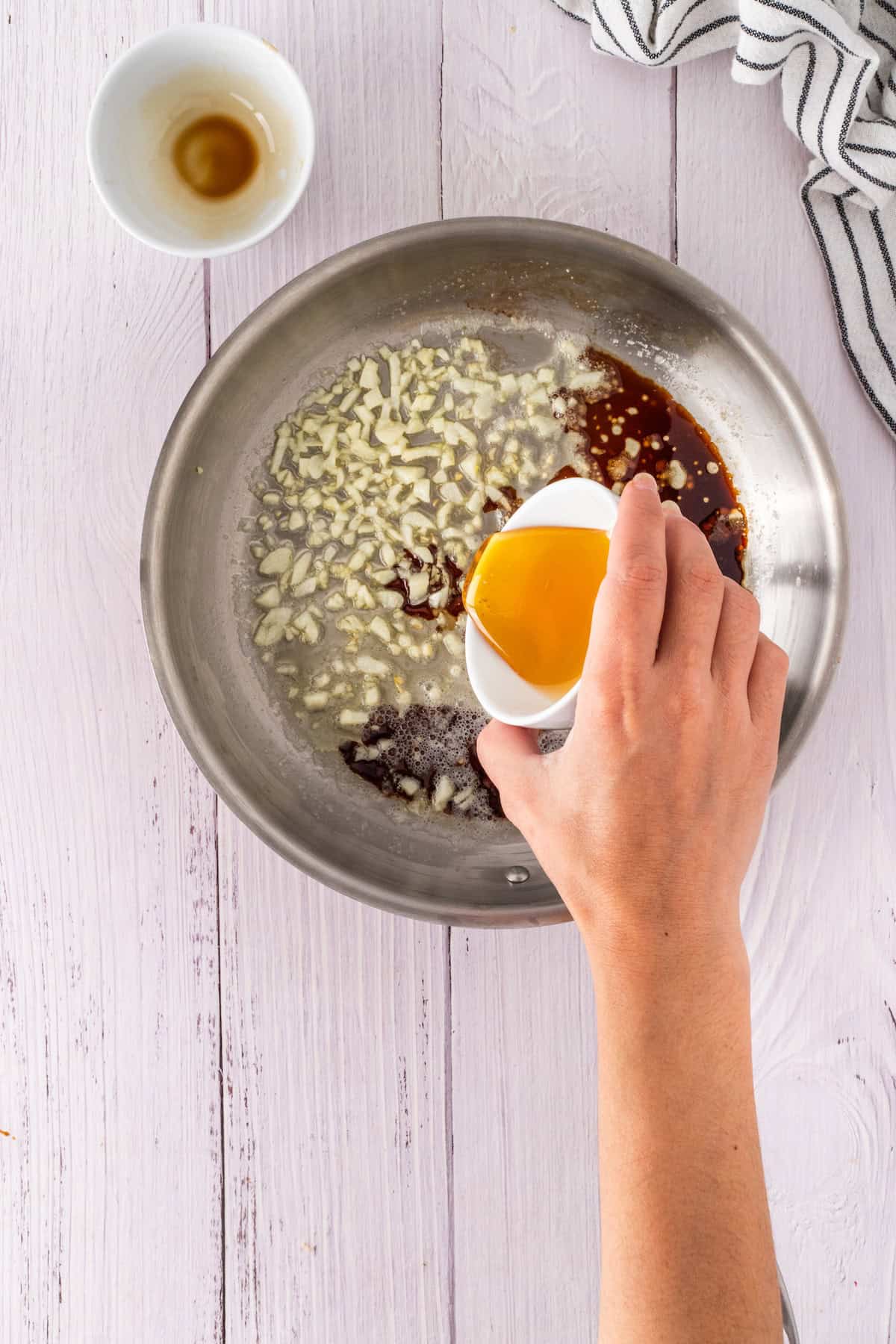 Hand pouring honey from a small white cup into a skillet with chopped garlic and a rich brown liquid. Striped cloth and empty cup sit nearby on the white wooden surface, ready for cooking magic.