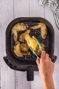 A hand holding green tongs lifts a crispy piece of fried chicken wings from an air fryer filled with delicious pieces. A white and black striped cloth is visible beside the air fryer, hinting at the perfect blend of honey and garlic seasoning.