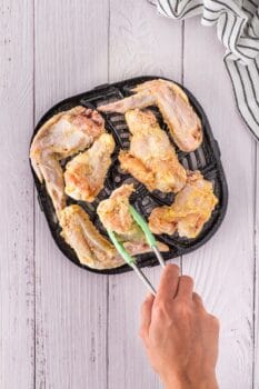 Raw chicken wings on a black grill pan with a hand using tongs to place one piece, set on a light wooden surface with a striped cloth nearby, exude an inviting aroma of garlic.