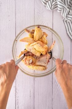 Hands tossing raw chicken wings glazed with honey and garlic in a glass bowl, on a white wooden surface with a striped cloth nearby.