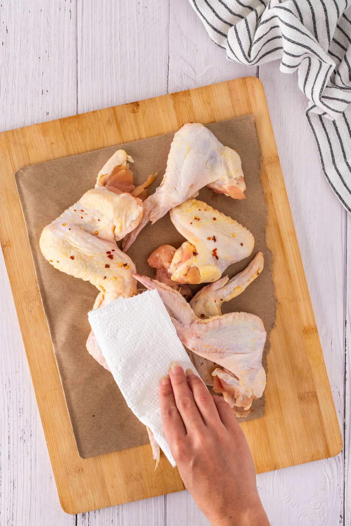 Person patting raw chicken wings dry with a paper towel on a wooden cutting board, preparing them for a delicious honey garlic glaze; white cloth nearby.