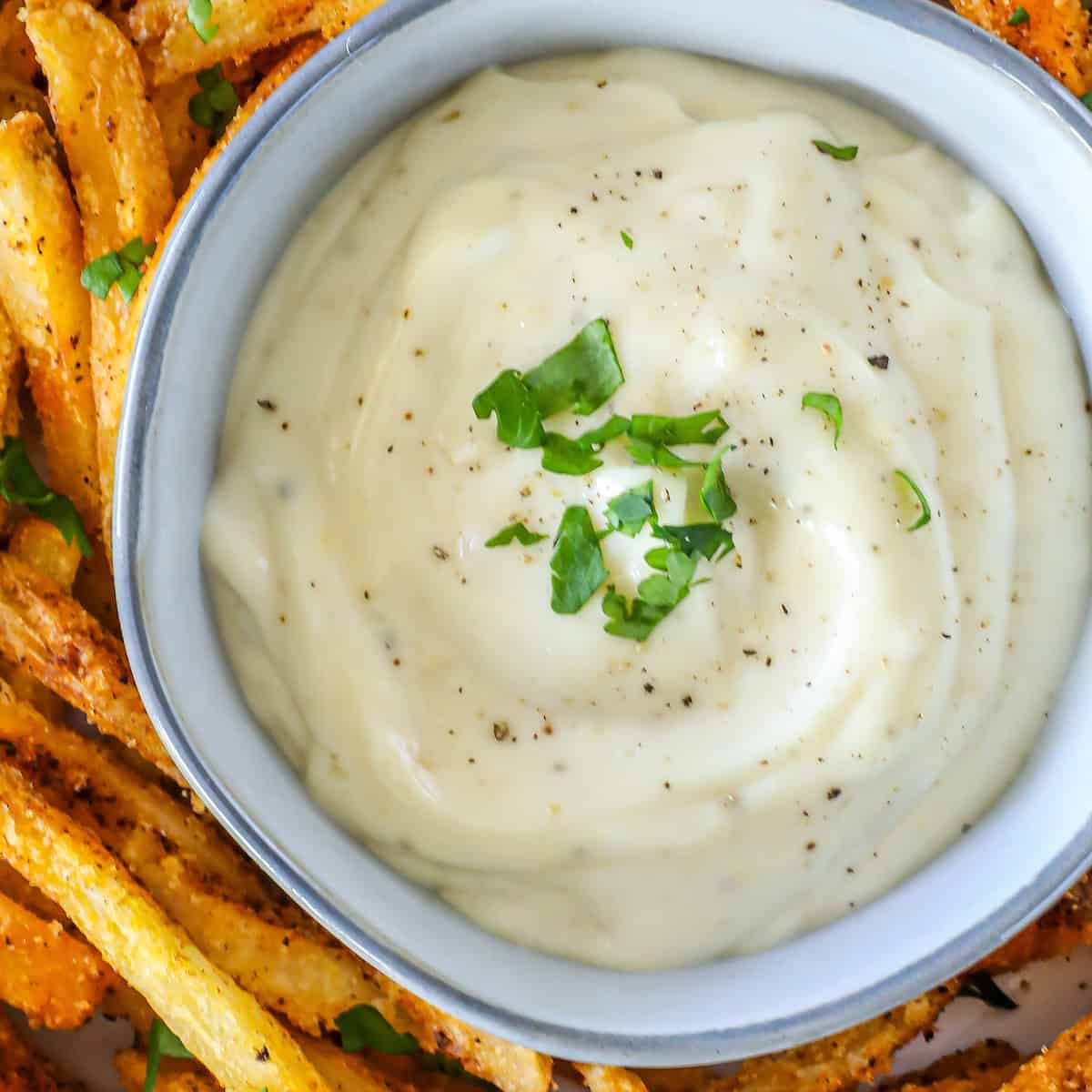 A bowl of creamy lemon garlic aioli garnished with black pepper and green herbs sits surrounded by crispy, seasoned French fries.