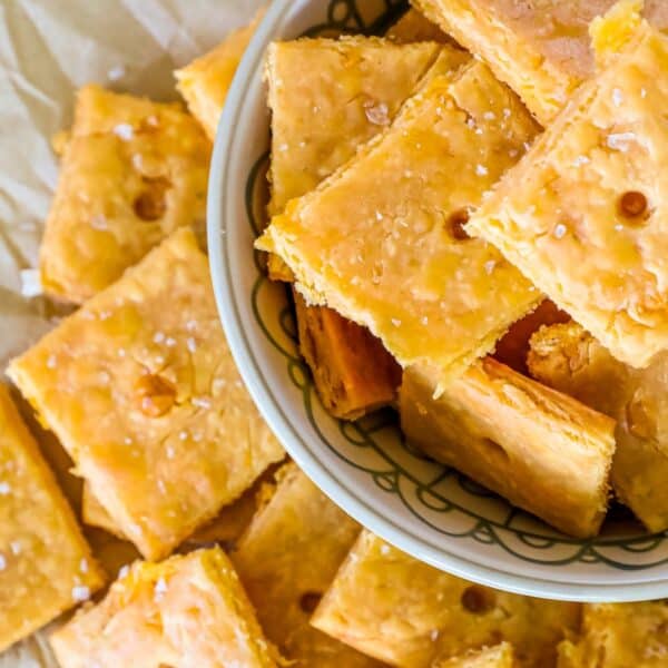 A bowl brimming with crispy, golden-brown sourdough crackers dusted with sea salt, surrounded by more on parchment paper.