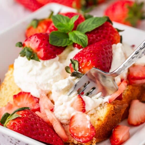 A fork in a slice of strawberry shortcake topped with whipped cream, fresh strawberries, and mint leaves on a white plate.