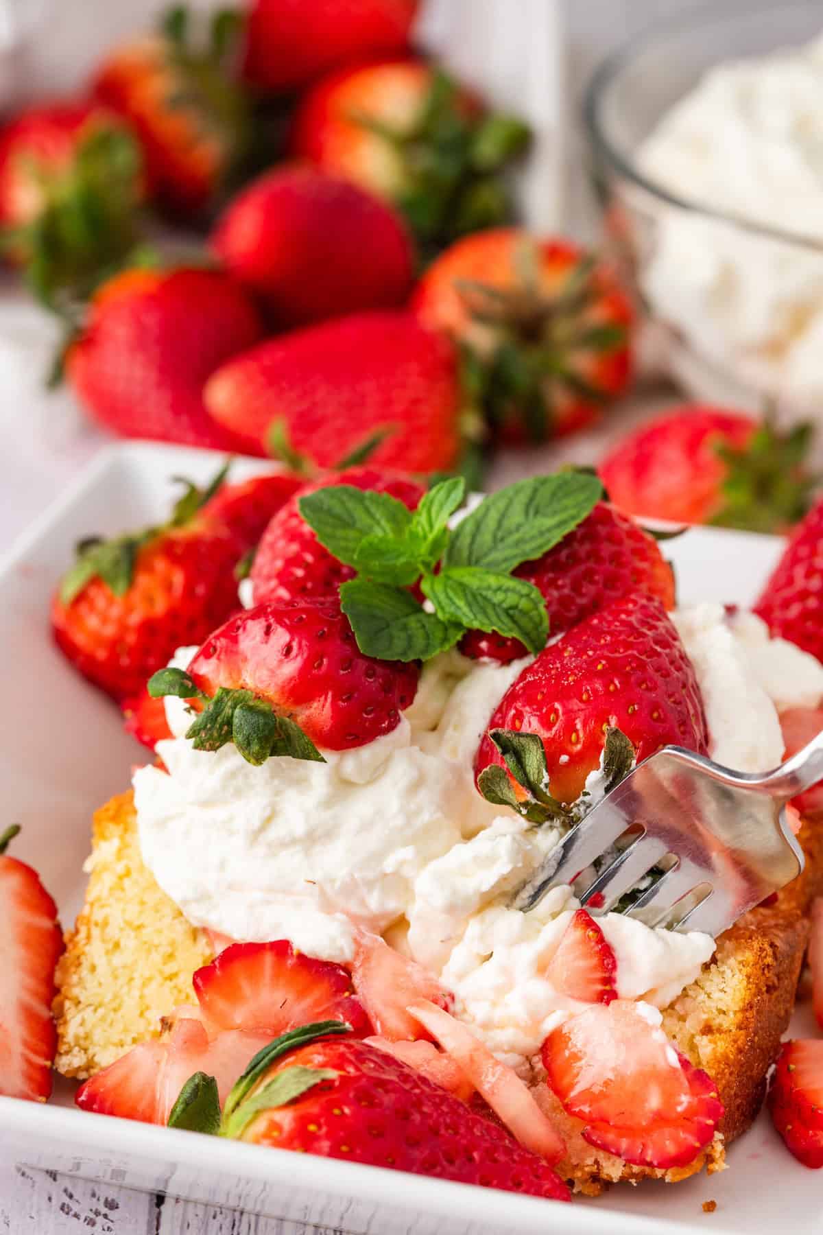 A slice of strawberry shortcake adorned with juicy strawberries, whipped cream, and a mint garnish rests on a white plate. A fork is cutting into the dessert. Fresh strawberries and a bowl of cream enhance the background's allure.