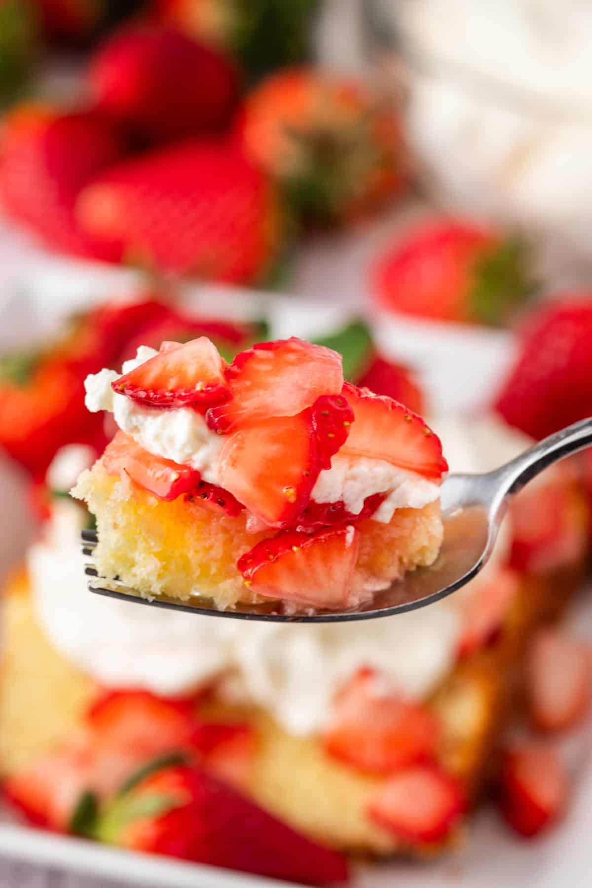 A close-up of a fork holding a bite of fluffy strawberry shortcake with whipped cream, framed by a larger helping and fresh strawberries in the background.