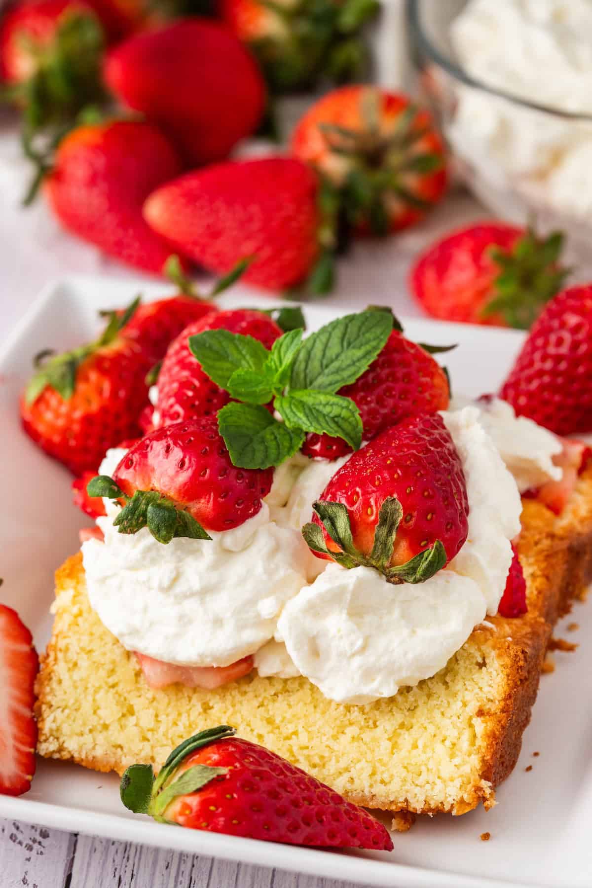 A slice of strawberry shortcake topped with whipped cream and fresh berries, garnished with a mint leaf on a white plate. Additional strawberries are artfully scattered around.