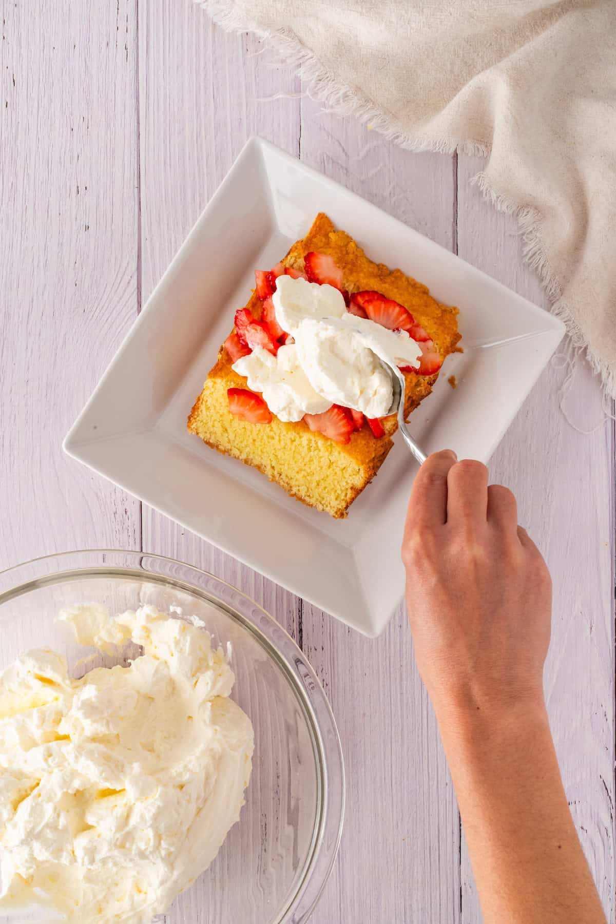 A hand adds whipped cream to a slice of strawberry shortcake on a square white plate, perfectly topped with fresh strawberries. A bowl of whipped cream sits beside it on the light wooden table.