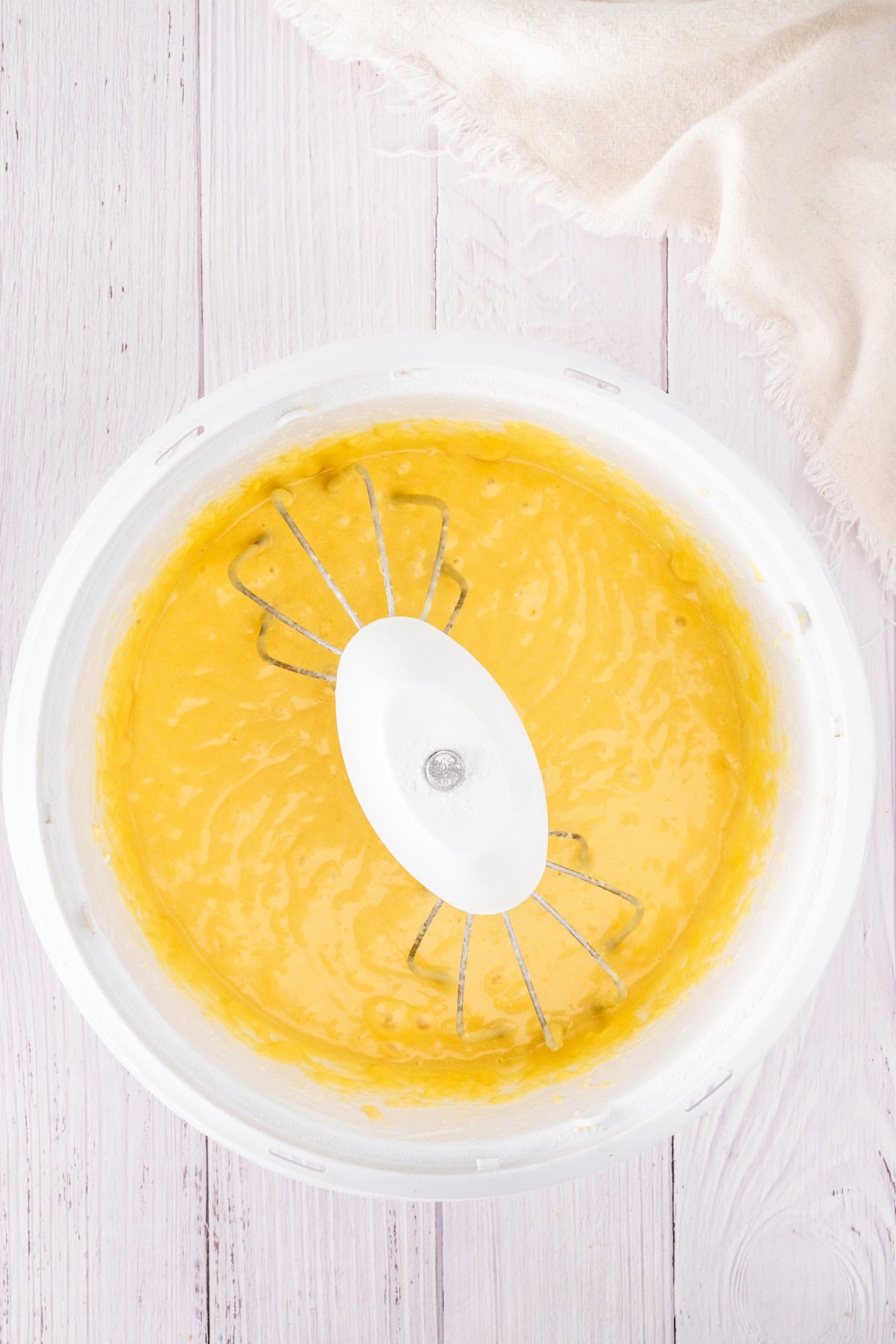 A bowl of yellow cake batter with a whisk attachment sits on a light wooden surface, hinting at the creation of a delightful strawberry shortcake. Nearby, a cloth rests casually, ready to assist in crafting this sweet treat.
