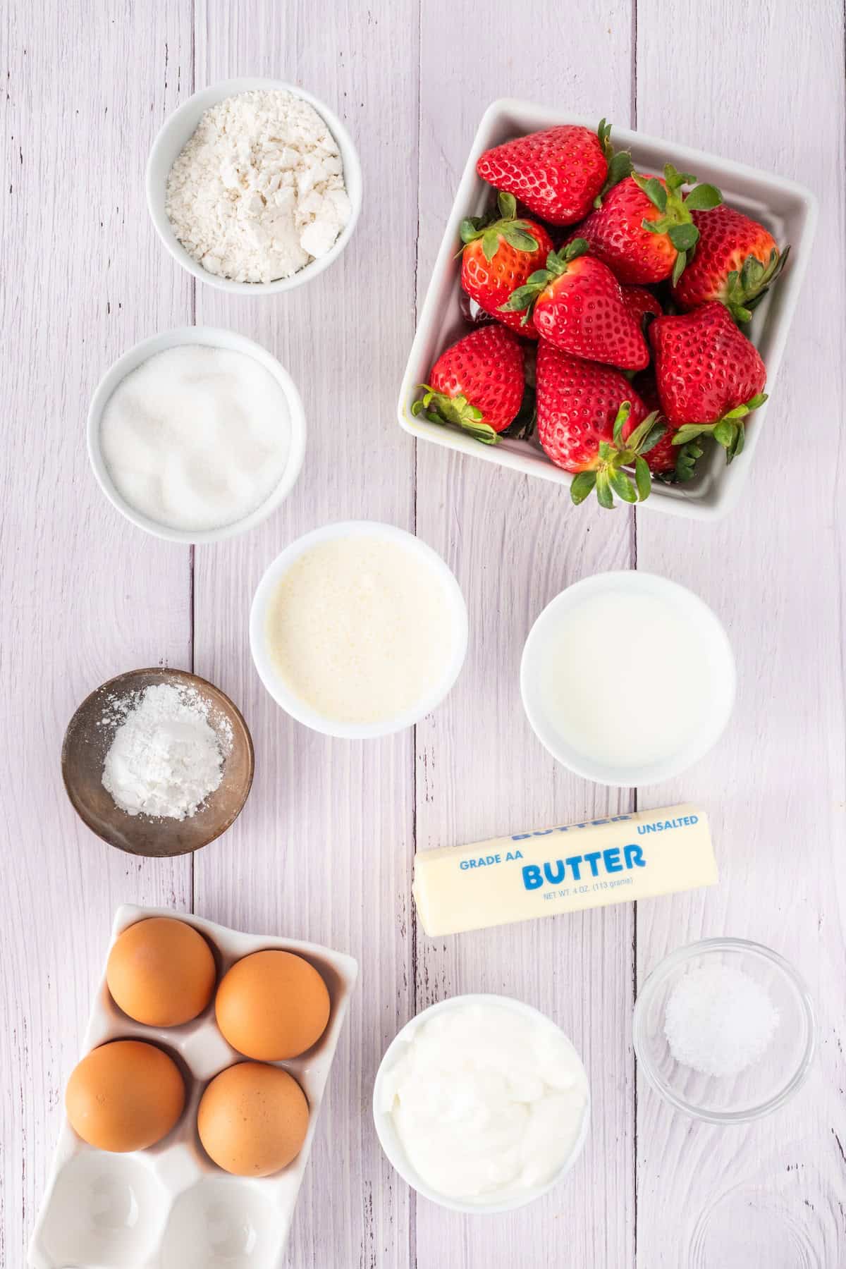 A delightful scene on a light wooden surface unveils the key ingredients for a strawberry shortcake: fresh strawberries, butter, flour, sugar, eggs, cream, yogurt, baking powder, and salt.