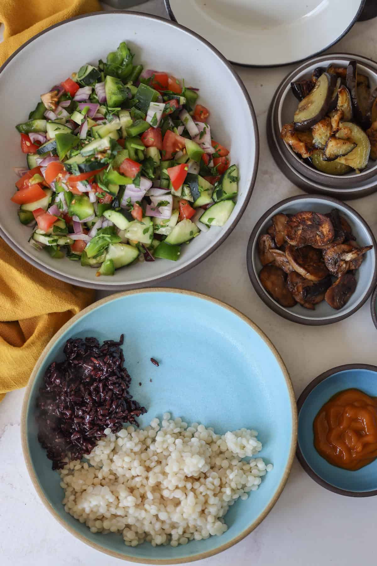 Bowls of vibrant mixed salad, grilled vegetables, couscous, and black rice accompany a small bowl of sauce on the table, creating a picturesque vegan feast reminiscent of a sabich spread.