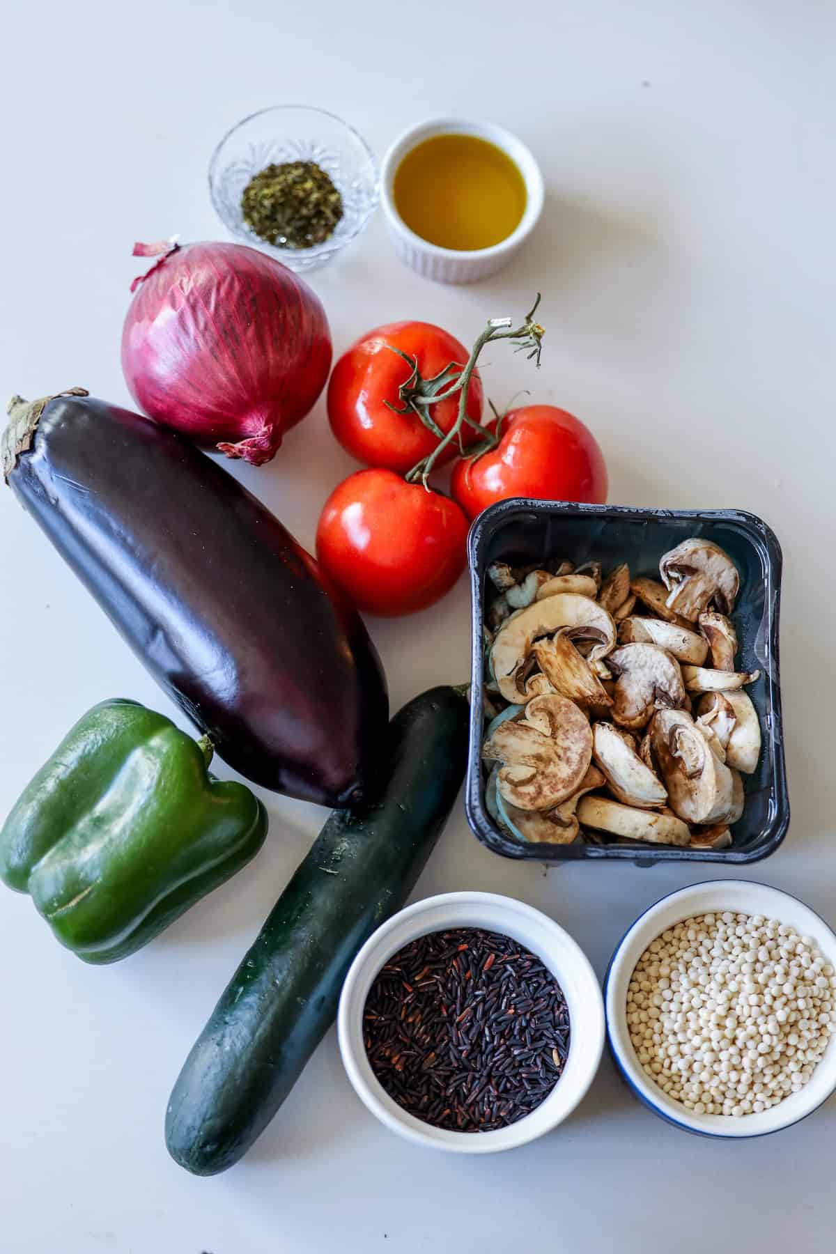 Assorted fresh vegetables including eggplant, red onion, tomatoes, green pepper, cucumber, and mushrooms elegantly arranged in bowls alongside olive oil, herbs, black rice, and couscous on a white surface. Perfect for creating a delightful vegan feast.
