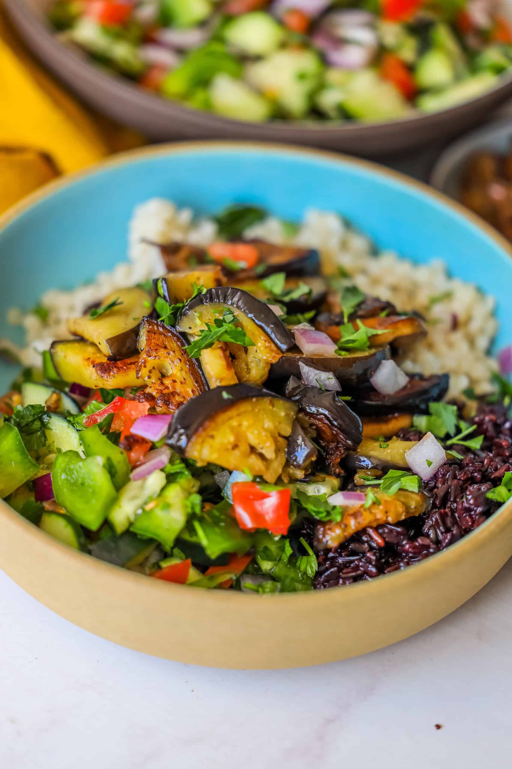 A bowl of mixed grains topped with roasted eggplant, diced tomatoes, cucumbers, and chopped herbs captures the essence of a vegan delight. Another bowl of Sabich-inspired ingredients sits partially visible in the background, enhancing the vibrant scene.
