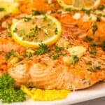Garlic Butter Salmon fillet grilled to perfection, garnished with lemon slices, chopped parsley, and garlic on a white plate.