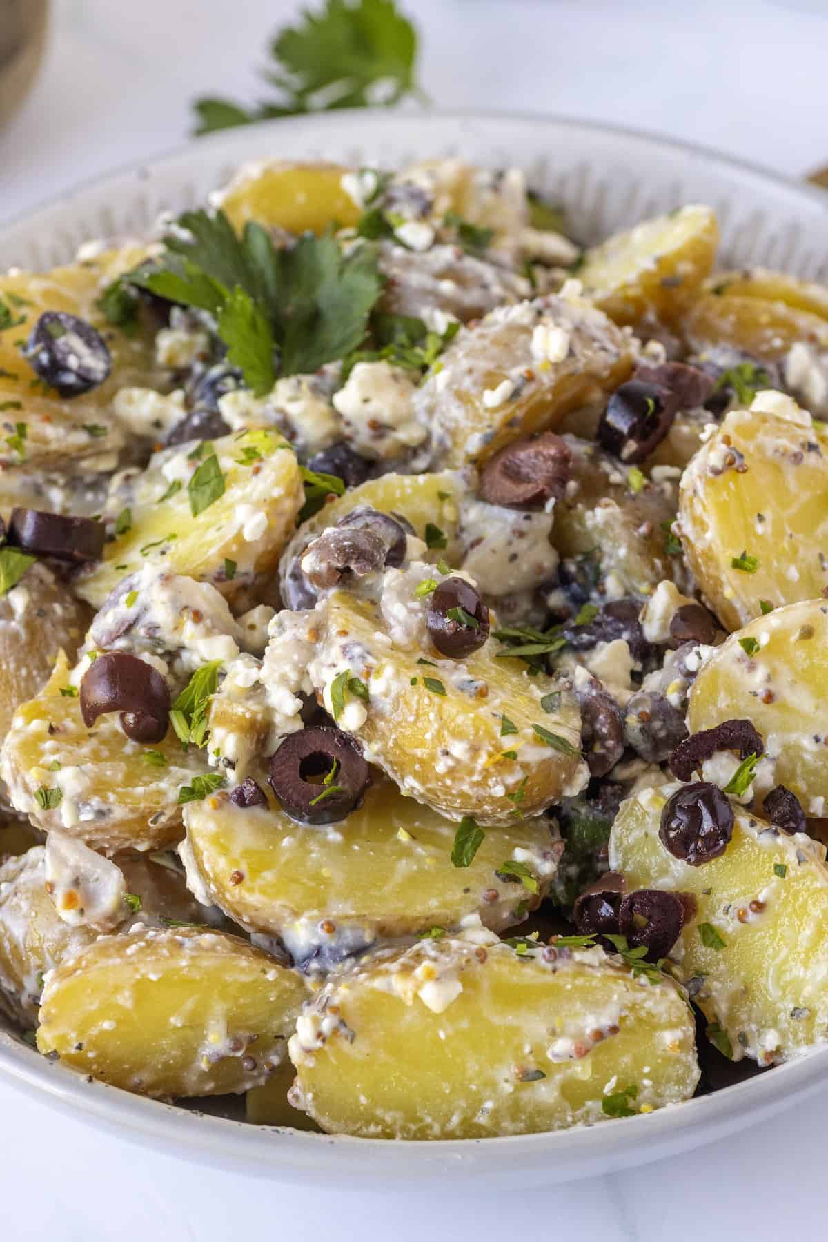 A bowl of Greek Potato Salad mixed with black olives, chopped herbs, and a creamy dressing.