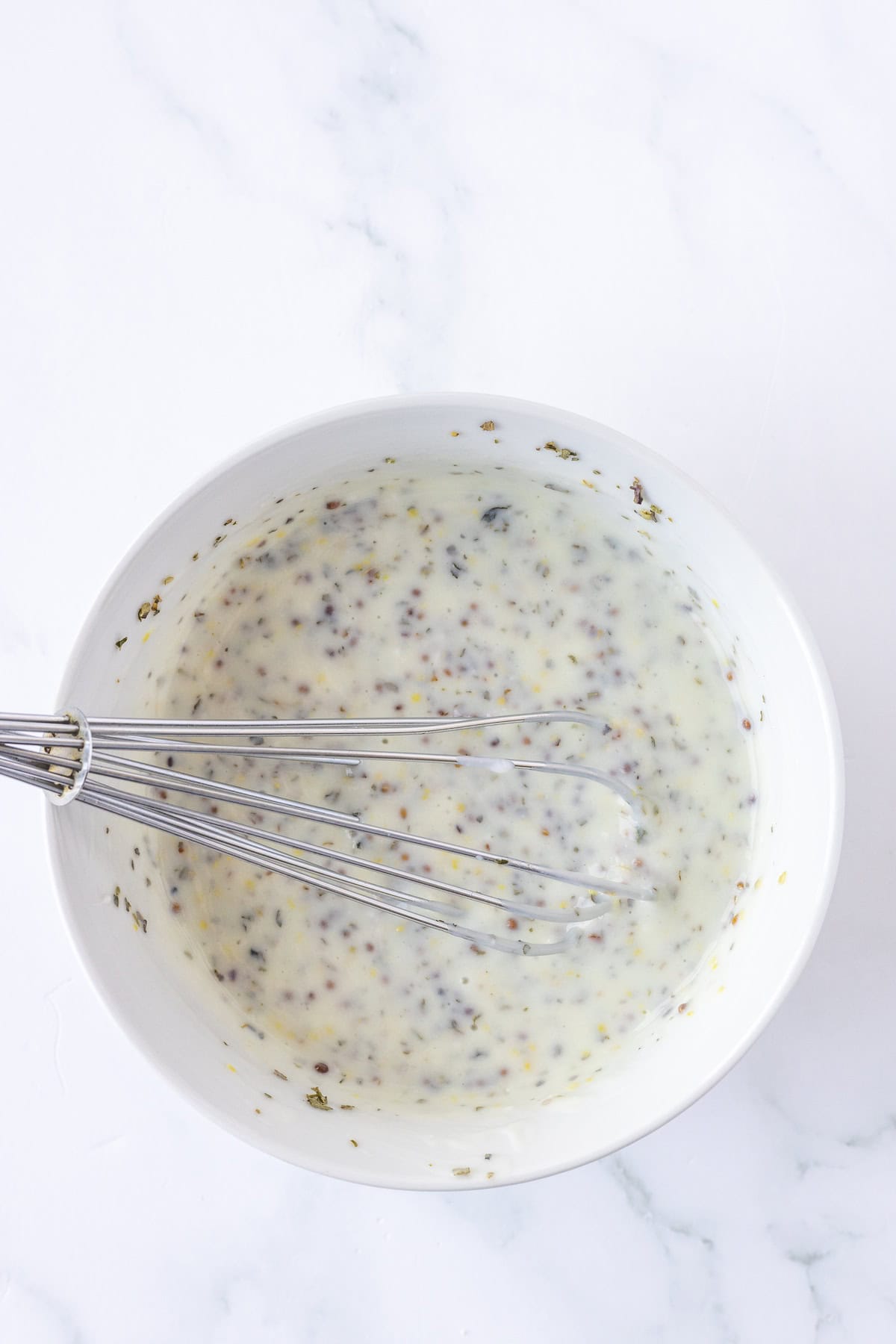 A metal whisk in a white bowl filled with creamy, speckled Greek Potato Salad dressing or sauce rests on a white marble surface.