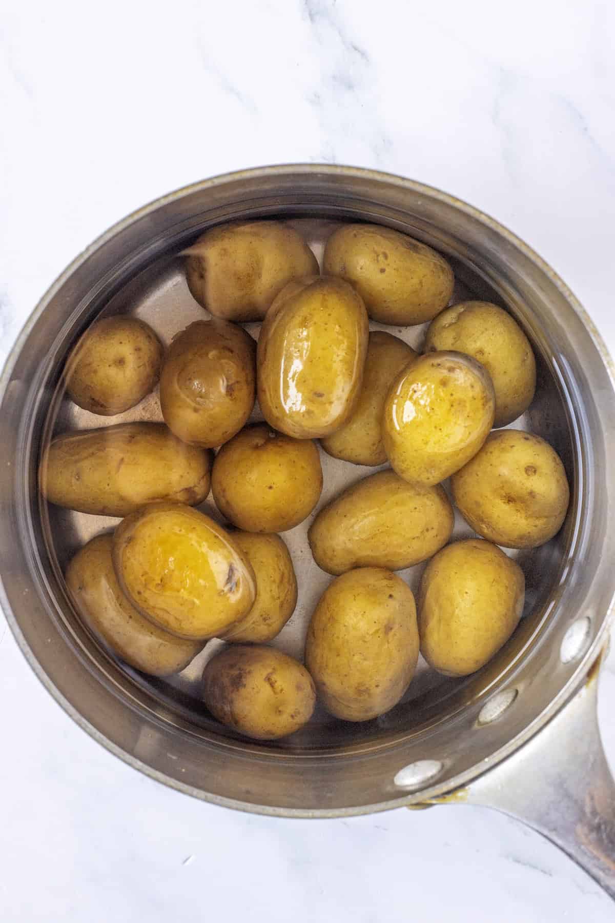 A stainless steel pot filled with whole, unpeeled potatoes submerged in water on a white marble surface, ready to be cooked for Greek Potato Salad.