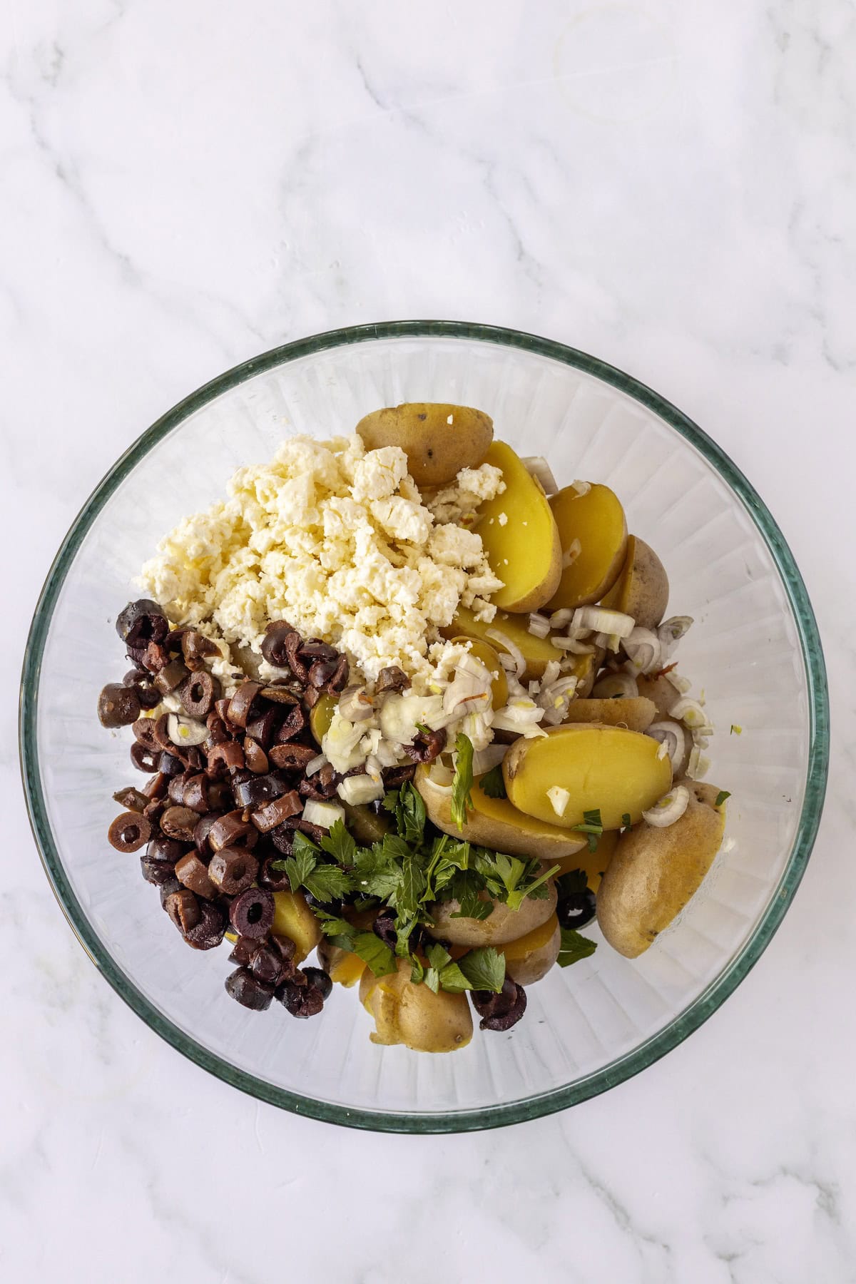 Glass bowl with sliced yellow potatoes, chopped black olives, crumbled feta cheese, sliced onions, and fresh parsley on a white marble surface—a vibrant Greek Potato Salad perfect for summer gatherings.