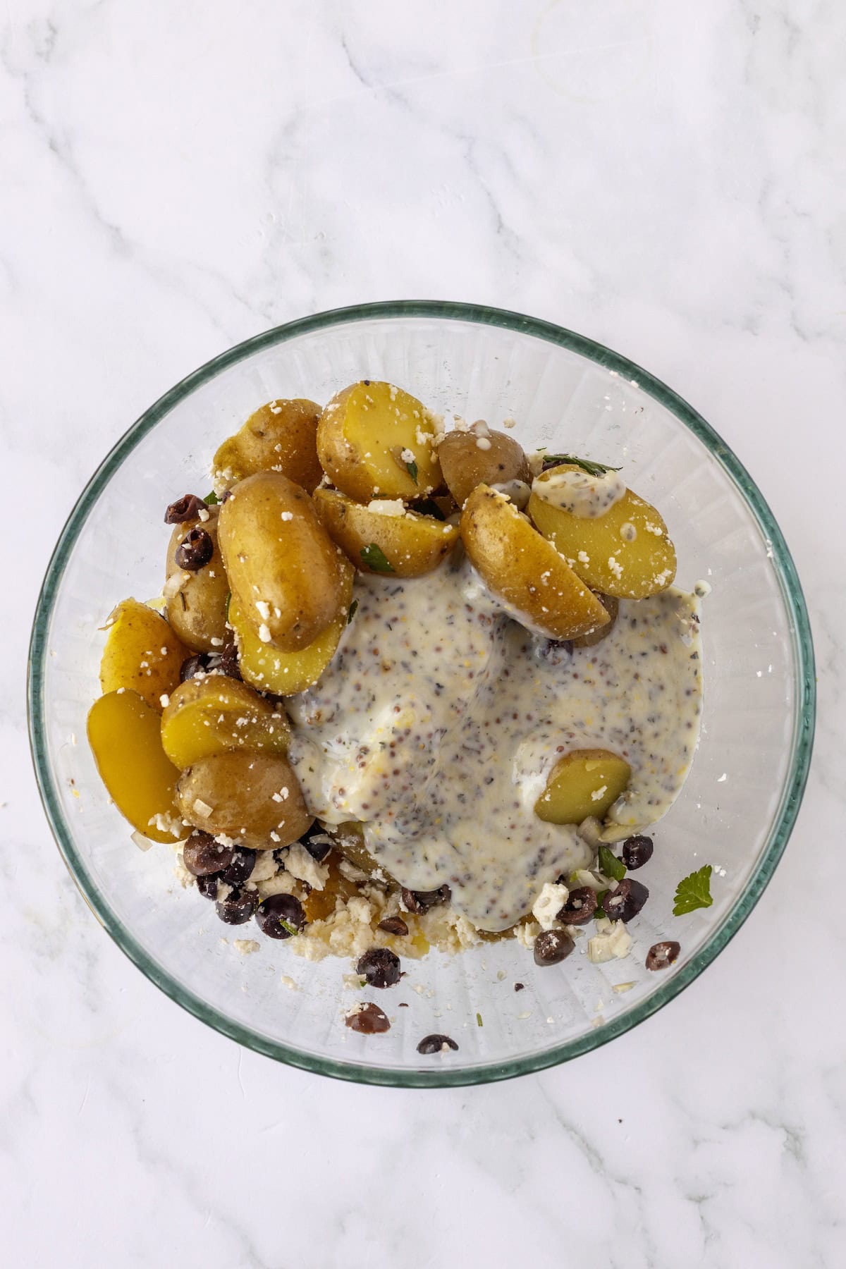 A bowl of Greek Potato Salad featuring halved baby potatoes, black beans, cheese crumbles, and a coarse-grain mustard dressing sits on a marble surface.