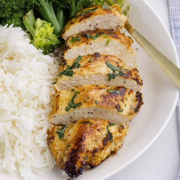 Sliced Baked Ranch Chicken breast garnished with herbs, served with white rice and steamed broccoli on a white plate.
