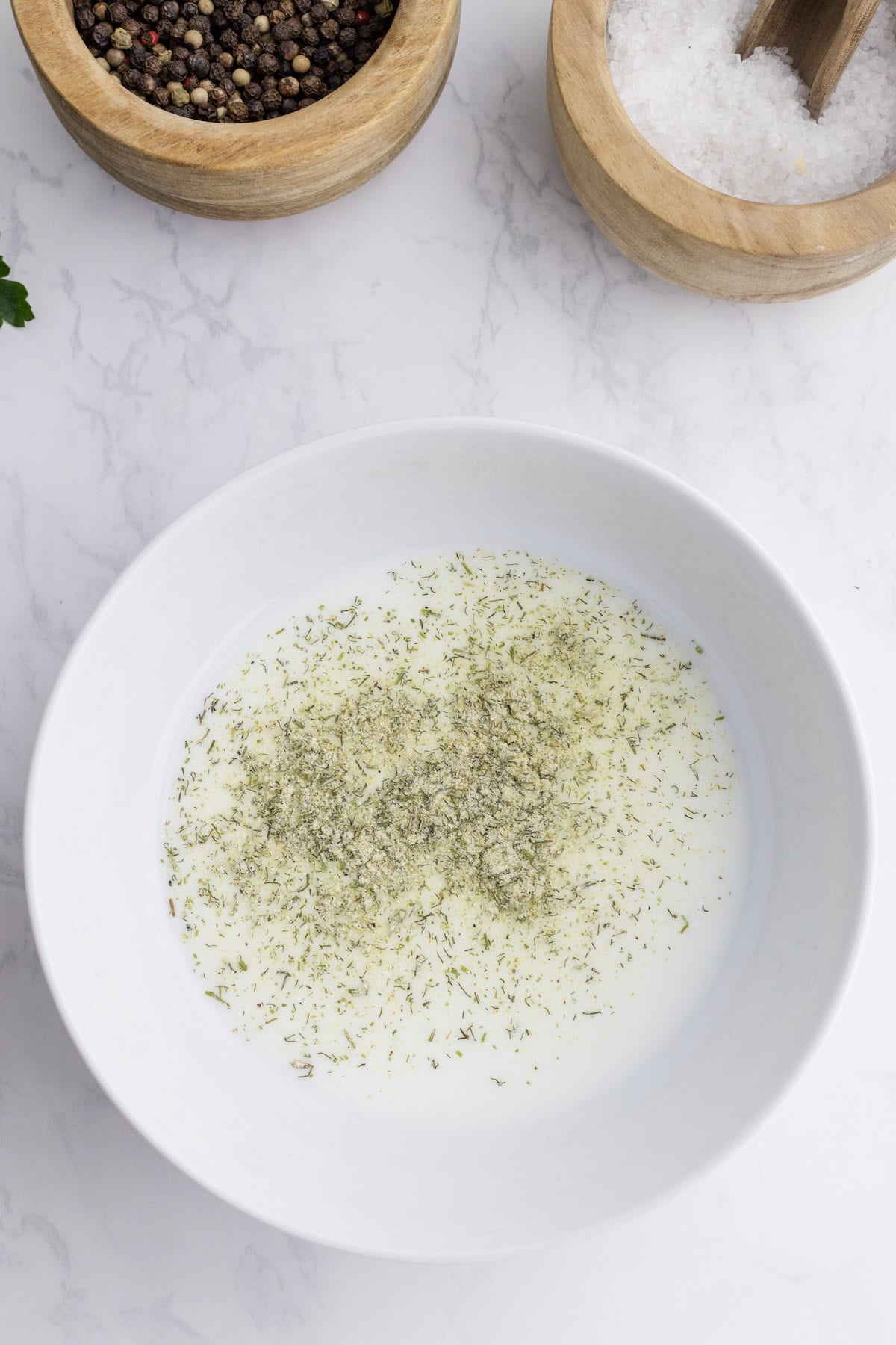 A white bowl with a creamy liquid and dried herbs, perfect for Ranch Chicken Breasts, placed on a marble surface next to bowls of peppercorns and salt.