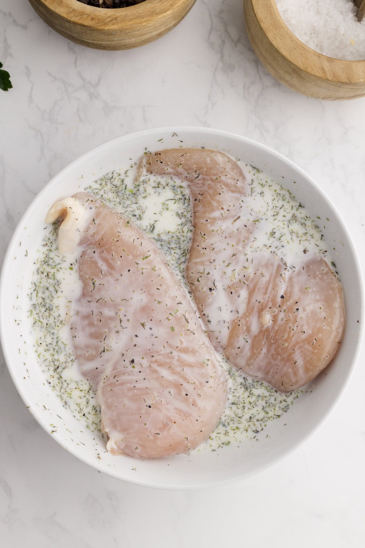 Raw Ranch Chicken Breasts marinating in a white bowl with herbs and a creamy liquid, set on a white marble surface beside wooden bowls of salt and spices.