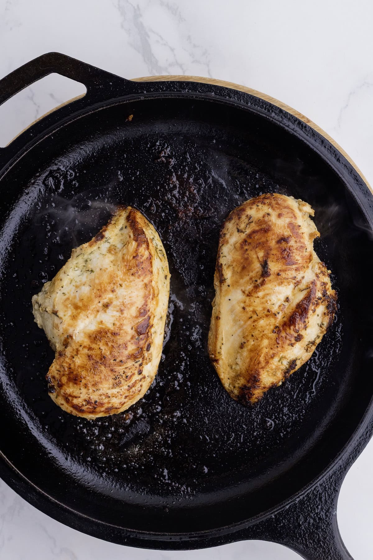 Two seasoned Ranch Chicken Breasts are cooked to perfection in a cast iron skillet, resting on a white marble surface—a simple yet flavorful addition to your favorite chicken recipes.