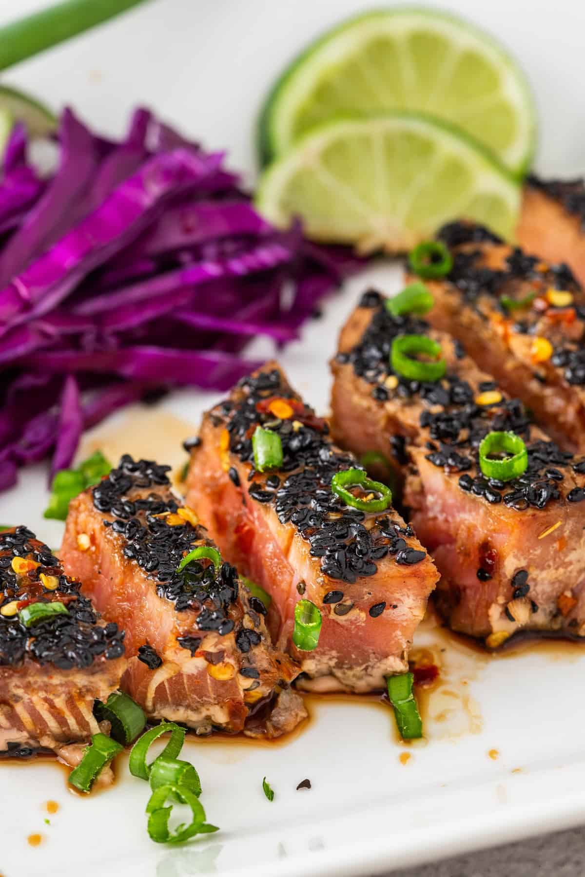 Seared Ahi Tuna, sliced and sesame-crusted, is garnished with green onions and served alongside shredded purple cabbage and lime slices on a white plate.