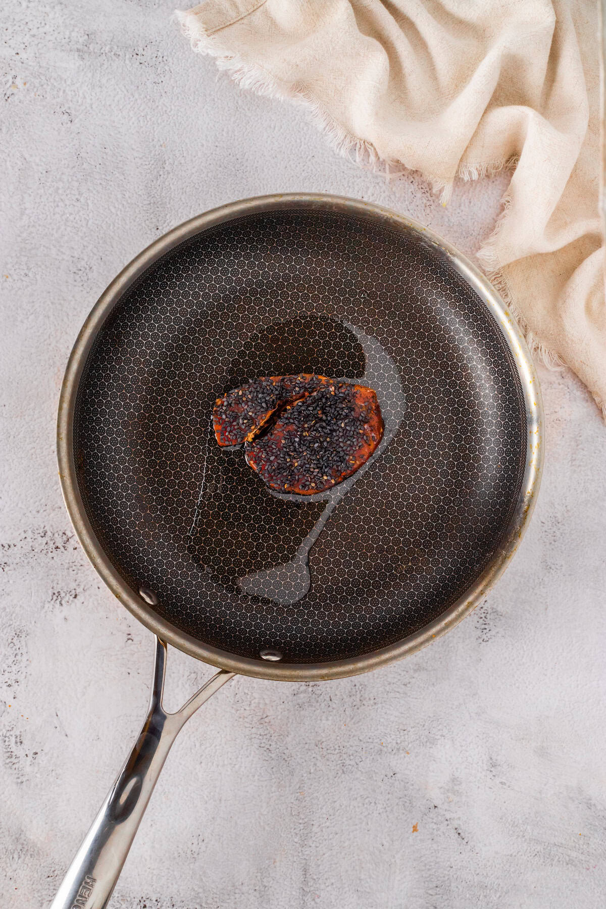 A burnt piece of food sits in the center of a frying pan with some oil, resembling overcooked Seared Ahi Tuna, on a light countertop next to a beige cloth.