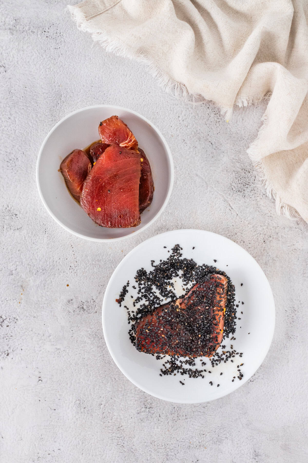 Two white plates on a light surface: one with slices of raw tuna, the other featuring Seared Ahi Tuna coated in black sesame seeds. A beige cloth is partially visible in the corner.