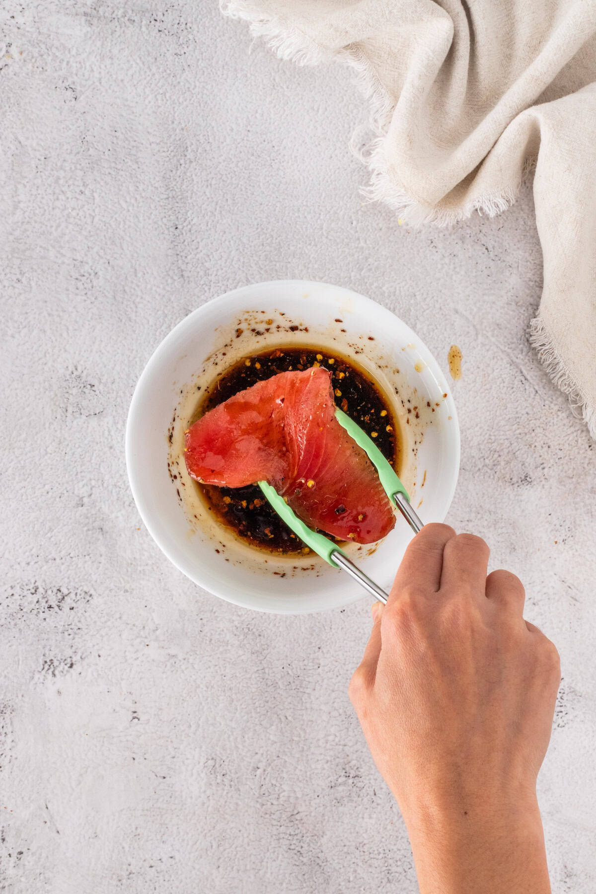 A hand uses tongs to dip a raw tuna steak into a bowl of soy-based marinade, preparing it for Seared Ahi Tuna, on a white surface with a beige cloth nearby.