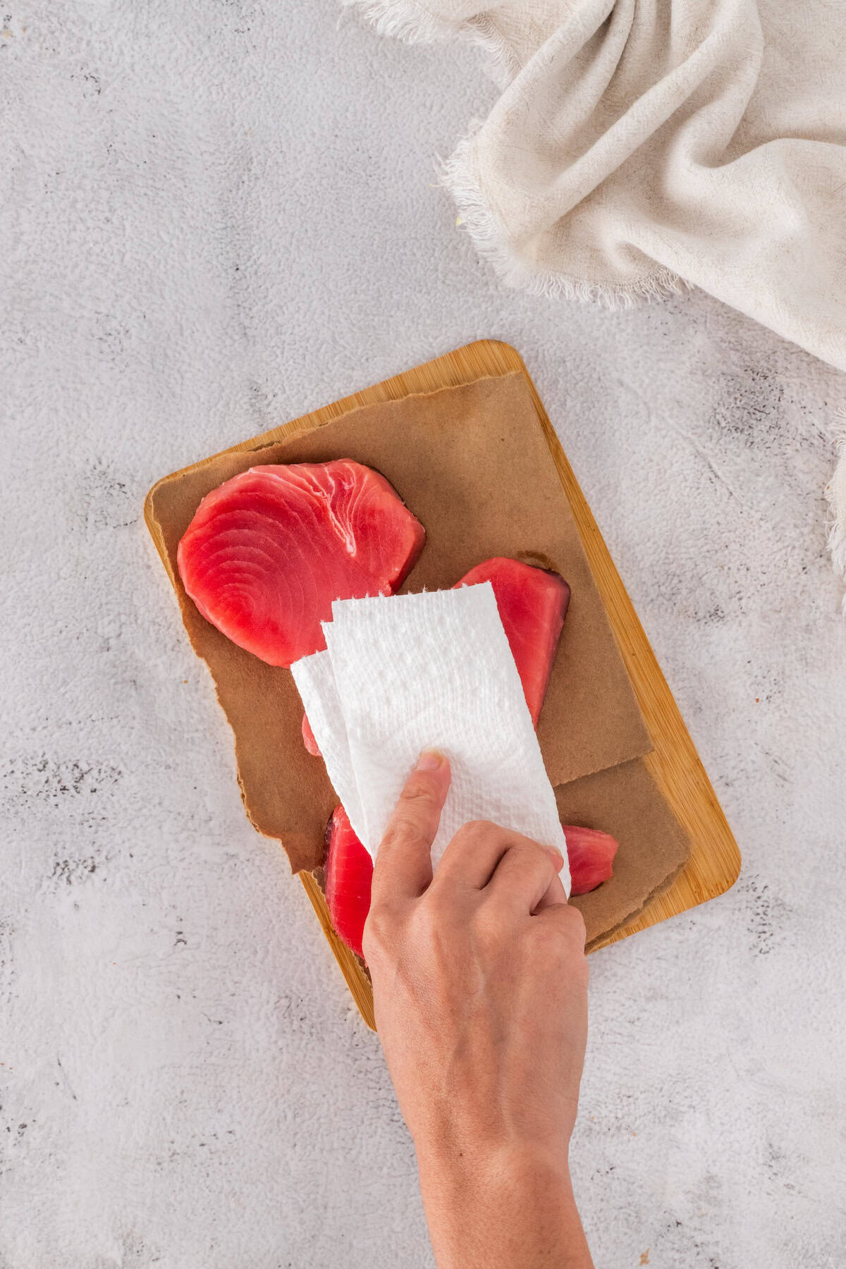 A hand uses a paper towel to pat dry raw tuna steaks for Seared Ahi Tuna on a wooden cutting board lined with parchment paper.