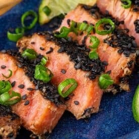 Sliced seared tuna topped with black sesame seeds, ginger, and chopped green onions, served on a blue plate with a slice of lime.
