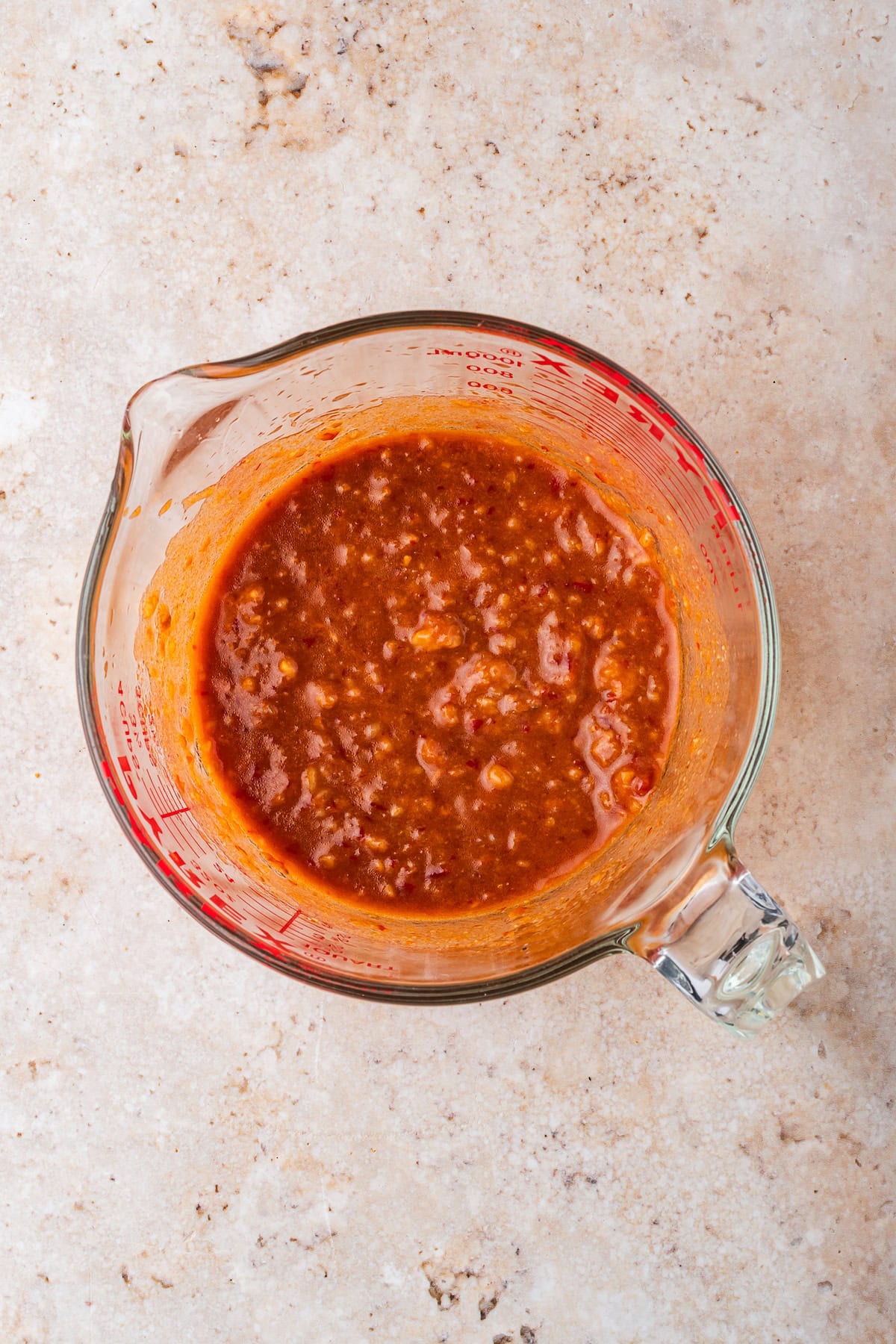 Glass measuring cup filled with chunky red sauce or salsa, perfect for topping your favorite Chicken Ramen or adding extra flavor to Slow Cooker recipes. Placed on a light-colored, textured surface.
