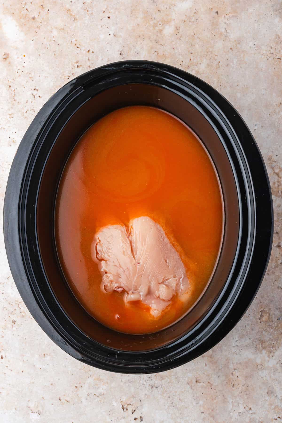 A raw chicken breast rests in orange-colored liquid inside a black slow cooker, perfect for preparing flavorful Slow Cooker Ramen, all set on a light brown countertop.