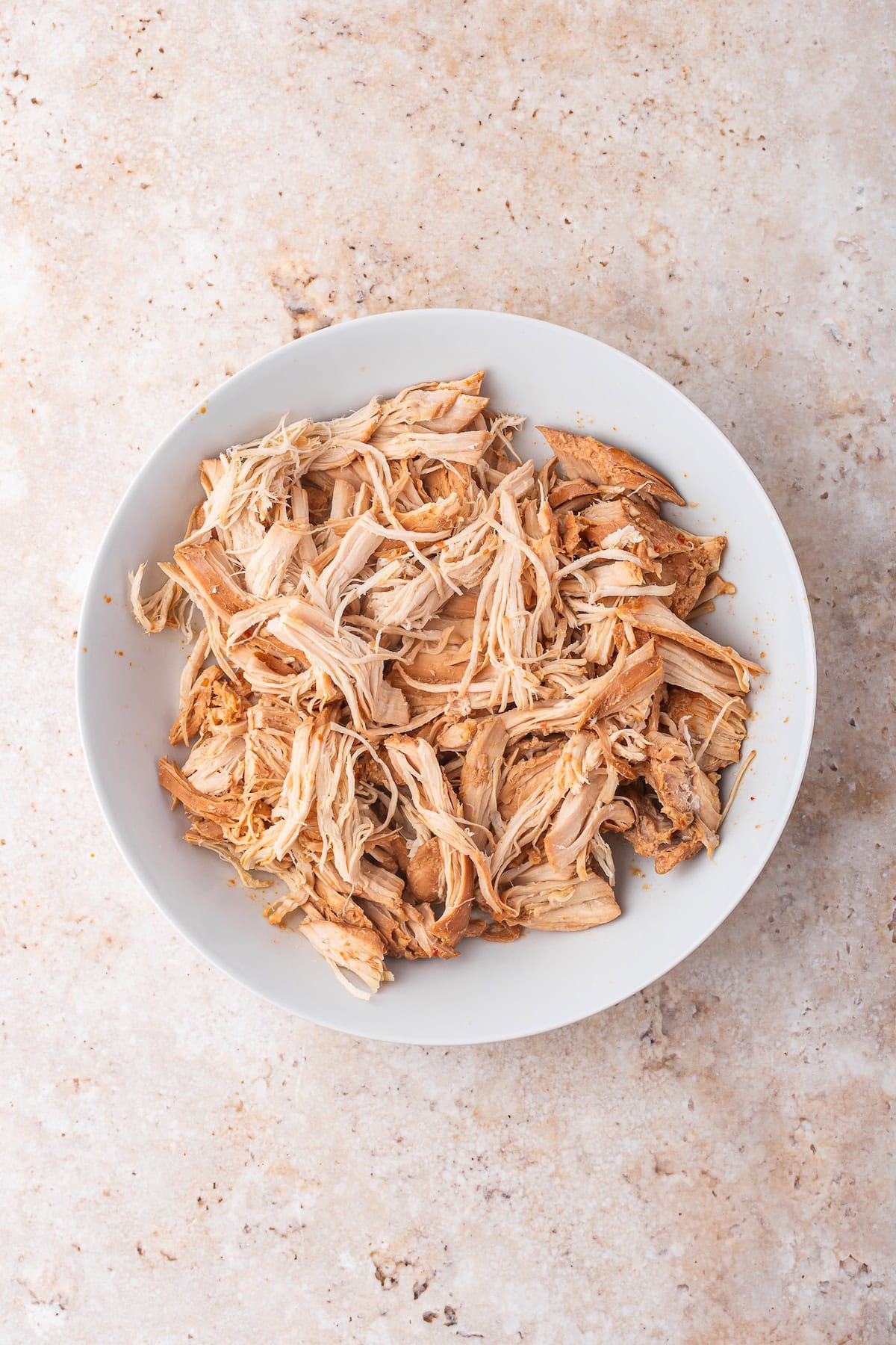A white bowl filled with shredded slow cooker chicken sits on a light-colored, textured surface—perfect for adding to your favorite chicken ramen.