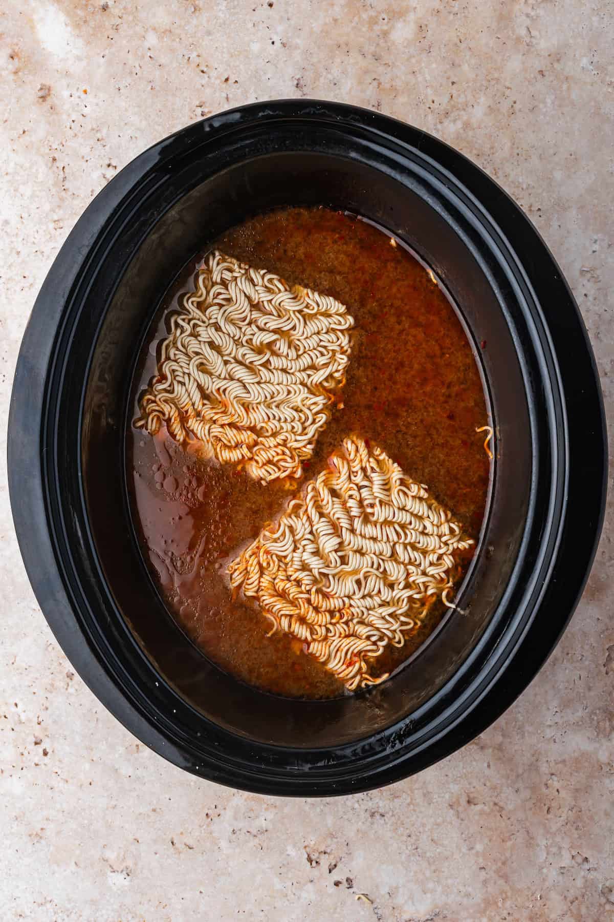 Two blocks of uncooked instant ramen noodles simmer in a slow cooker with savory broth, creating an easy and comforting Chicken Ramen meal.