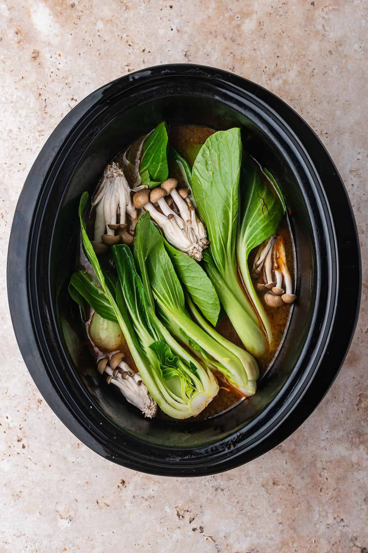 A slow cooker filled with bok choy, mushrooms, and broth for Slow Cooker Chicken Ramen, viewed from above on a light stone surface.