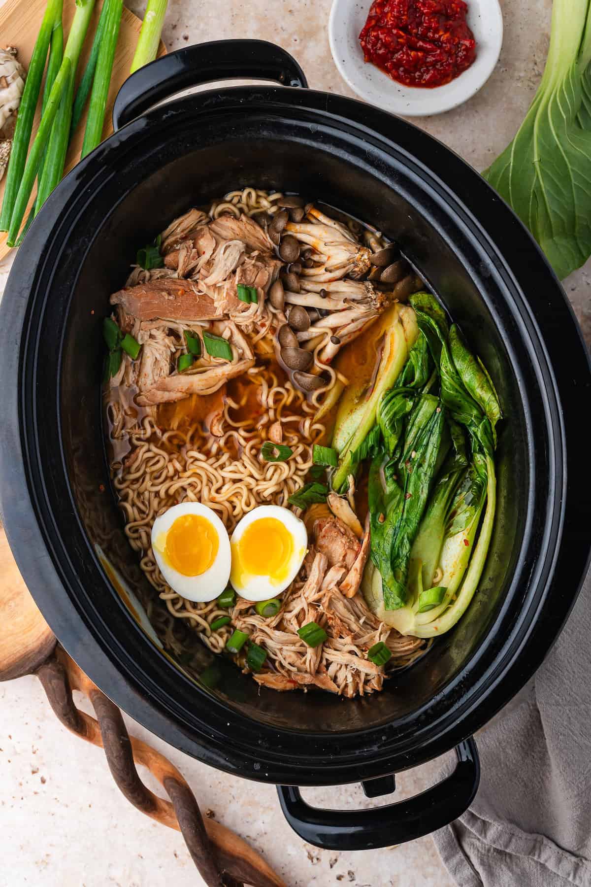 A bowl of Slow Cooker Ramen in a black pot with noodles, shredded chicken, soft-boiled eggs, mushrooms, and bok choy, garnished with green onions.