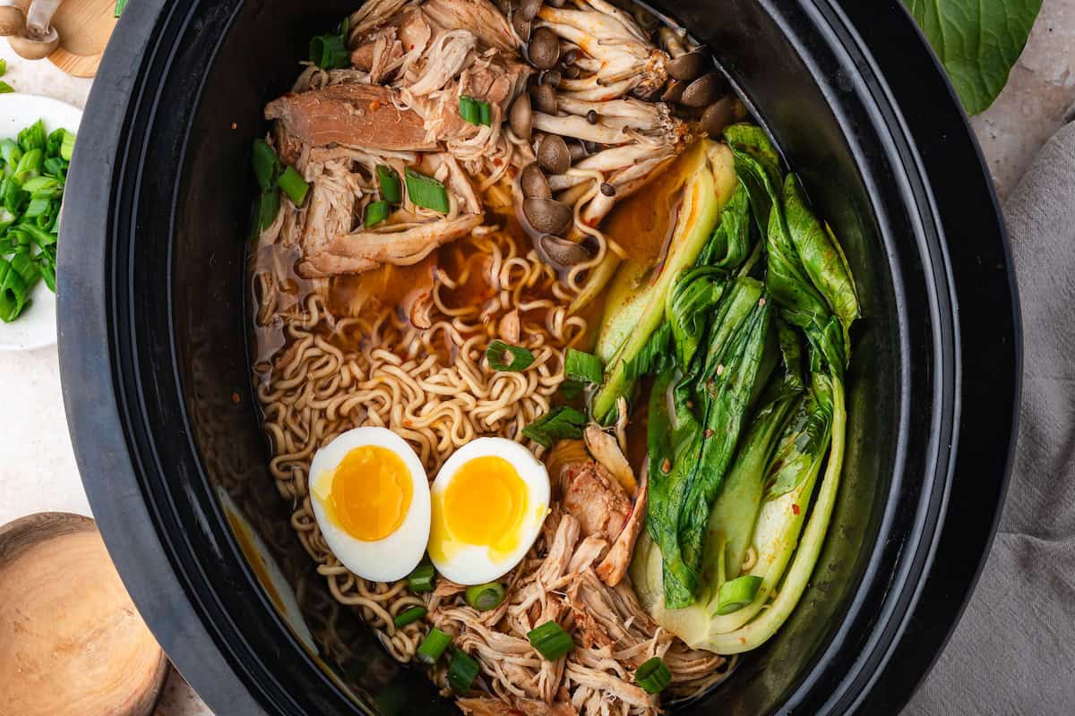 A slow cooker filled with savory chicken ramen, featuring shredded slow cooker chicken, bok choy, mushrooms, chopped green onions, ramen noodles, and two halved soft-boiled eggs in a flavorful broth.