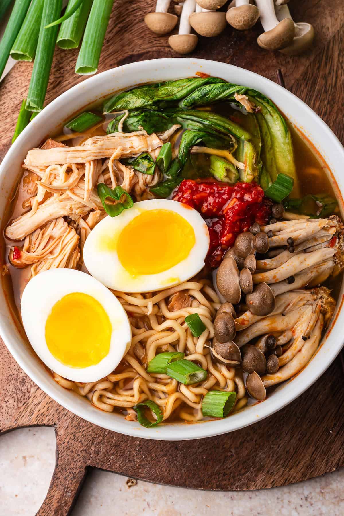 A bowl of Slow Cooker Chicken Ramen topped with soft-boiled eggs, shredded chicken, mushrooms, greens, scallions, and chili paste sits on a wooden board with green onions and mushrooms nearby.