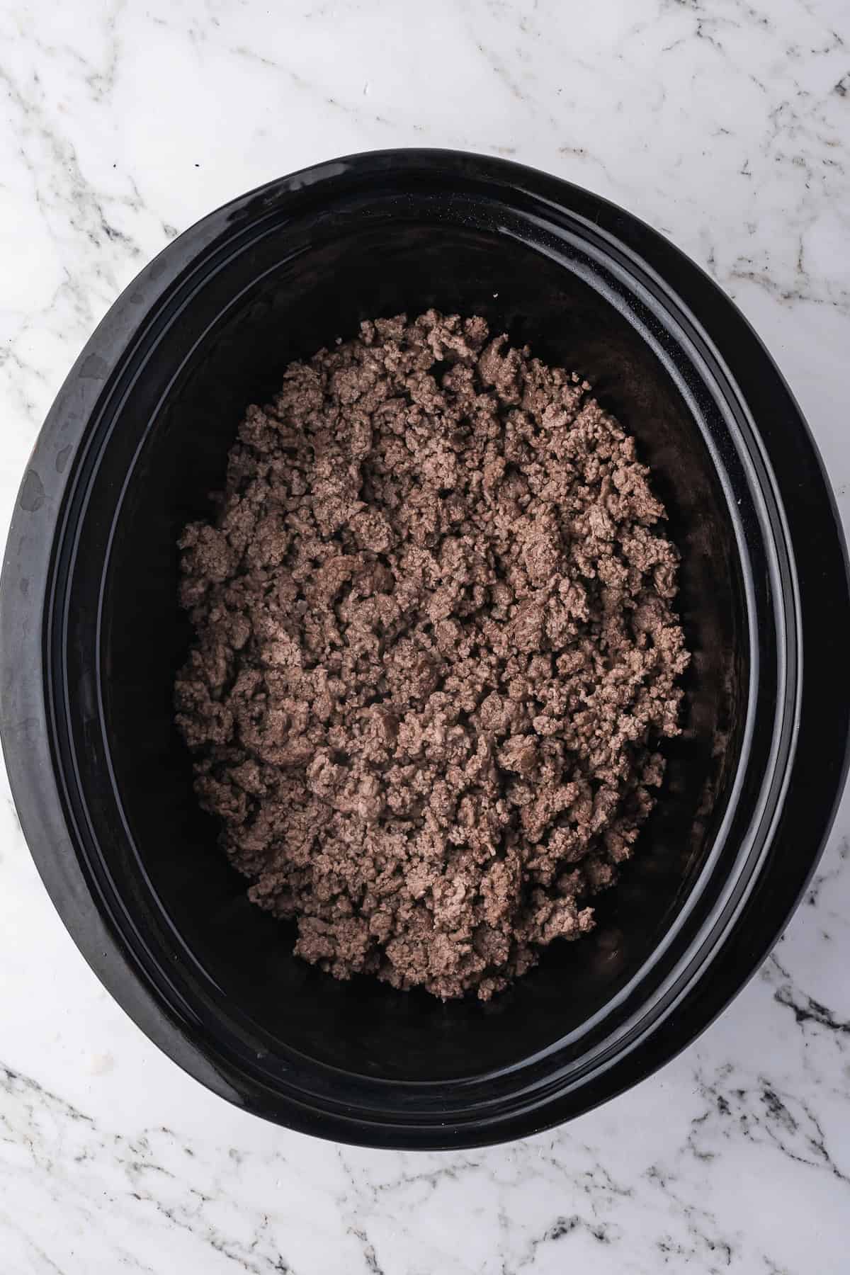 Tender cooked ground beef in a black slow cooker sits on a white marble surface—perfect for adding to your favorite beef recipes like classic Beef Stroganoff.