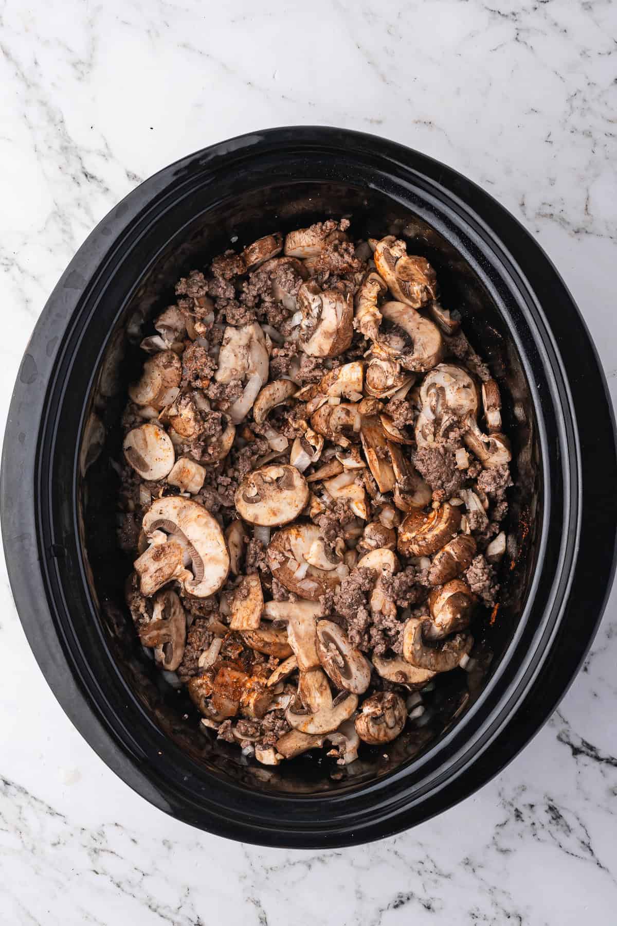 A black slow cooker filled with sliced mushrooms and ground beef sits on a white marble surface, perfect for making classic Beef Stroganoff or other hearty beef recipes.