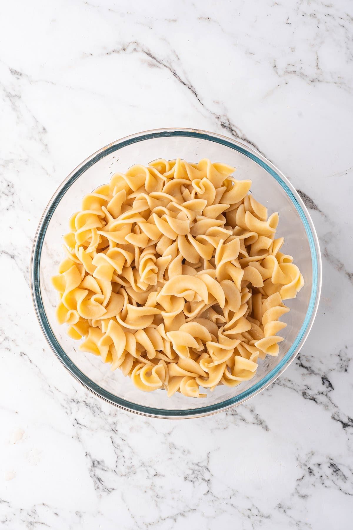 A glass bowl filled with plain, cooked egg noodles sits on a white marble surface, ready to be topped with your favorite Beef Stroganoff or slow cooker beef recipe.